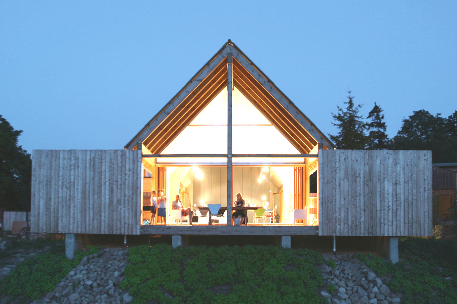Haus Michael – Das Haus mit dem Dreiecksgiebel steht auf einem Grundstück am See. Das Einfamilienhaus aus Lärchenholz teilt sich in 90 Quadratmeter Wohnfläche und eine Terrasse mit einer Fläche von 120 Quadratmetern auf. Grosse Schiebetüren an beiden Seiten des Hauses bilden die Eingänge. Durch einen Dachüberstand entsteht eine grosse Loggia, eine traufhohe Wand umschliesst wie ein Zaun das Haus.