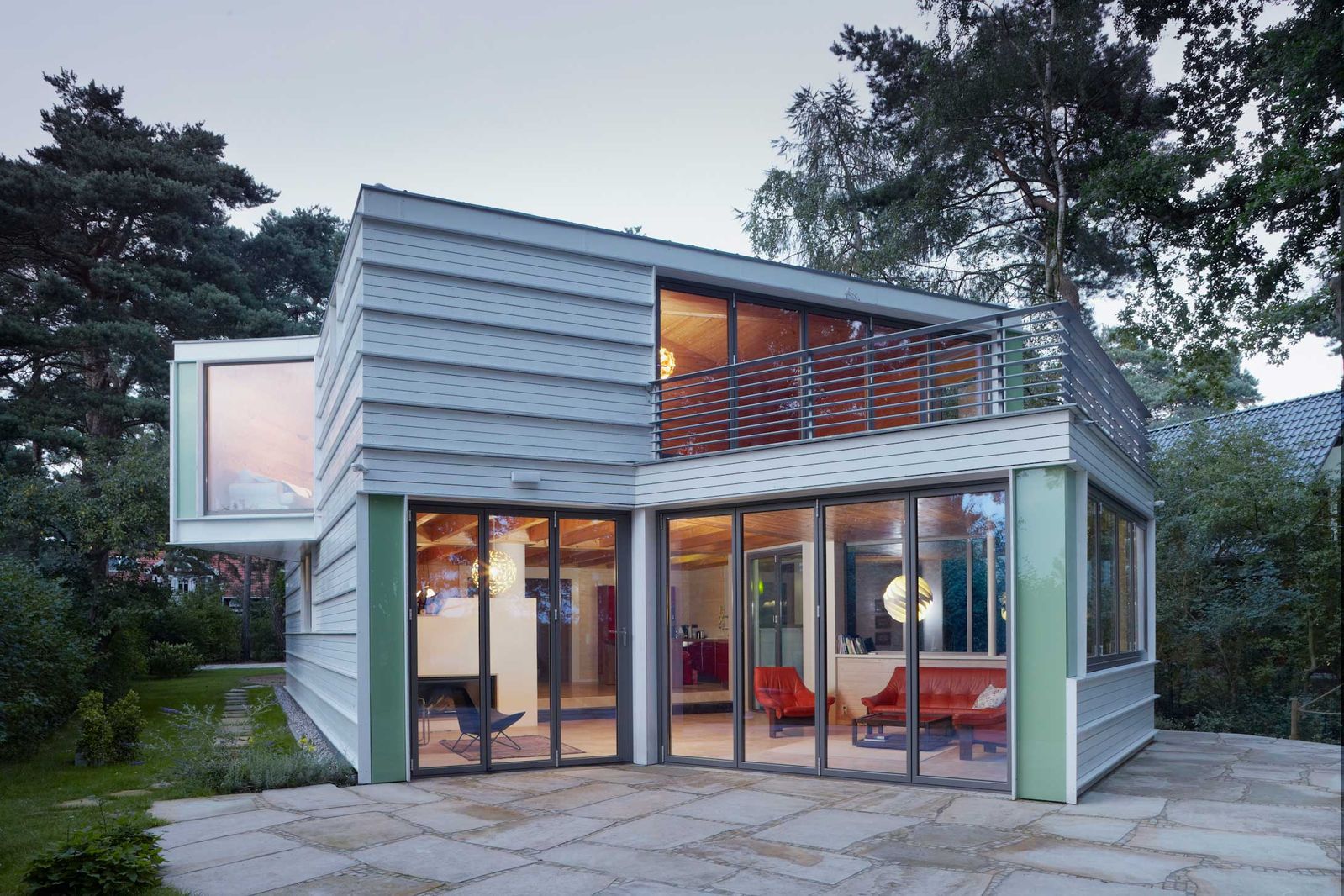 Holzhaus am See – Die weisse Farbe lässt das Holzhaus am See in kubischer Form und Massivbauweise gross und ausladend wirken. Das Haus überzeugt mit einer modernen, futuristisch anmutenden Form und einer Wohnfläche von 240 Quadratmetern. Als Raumtrenner dient ein eingebauter Kamin, der im Winter für angenehme Wärme sorgt. Ein dreiseitig verglaster Wintergarten bietet einen schönen Blick auf die weiträumige Terrasse und den See.