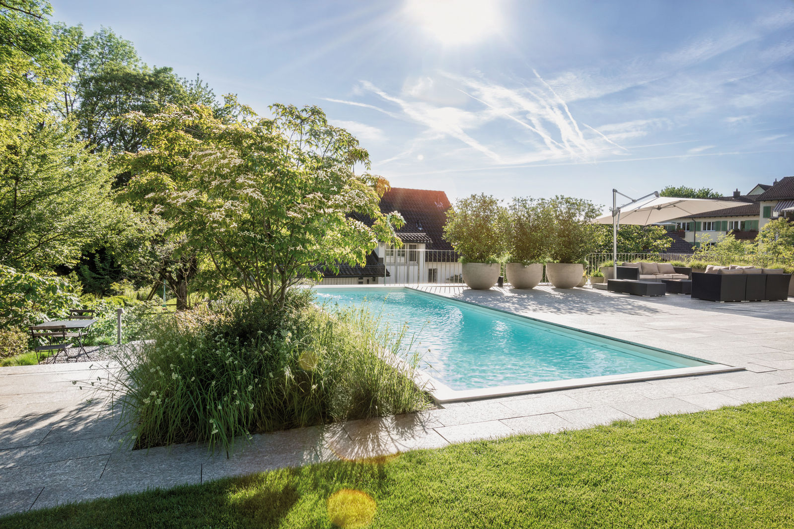 Swimmingpool im Familiengarten: Der Pool zieht alle Blicke auf sich. Eine grosse, mit Natursteinplatten verlegte Terrasse lädt zum Verweilen ein, die Rasenfläche wird von einer Sitzbank und zwei Gefässen eingerahmt. Im hinteren Bereich liegt leicht erhöht ein zweiter Sitzplatz, ebenfalls mit Blick auf den Pool.