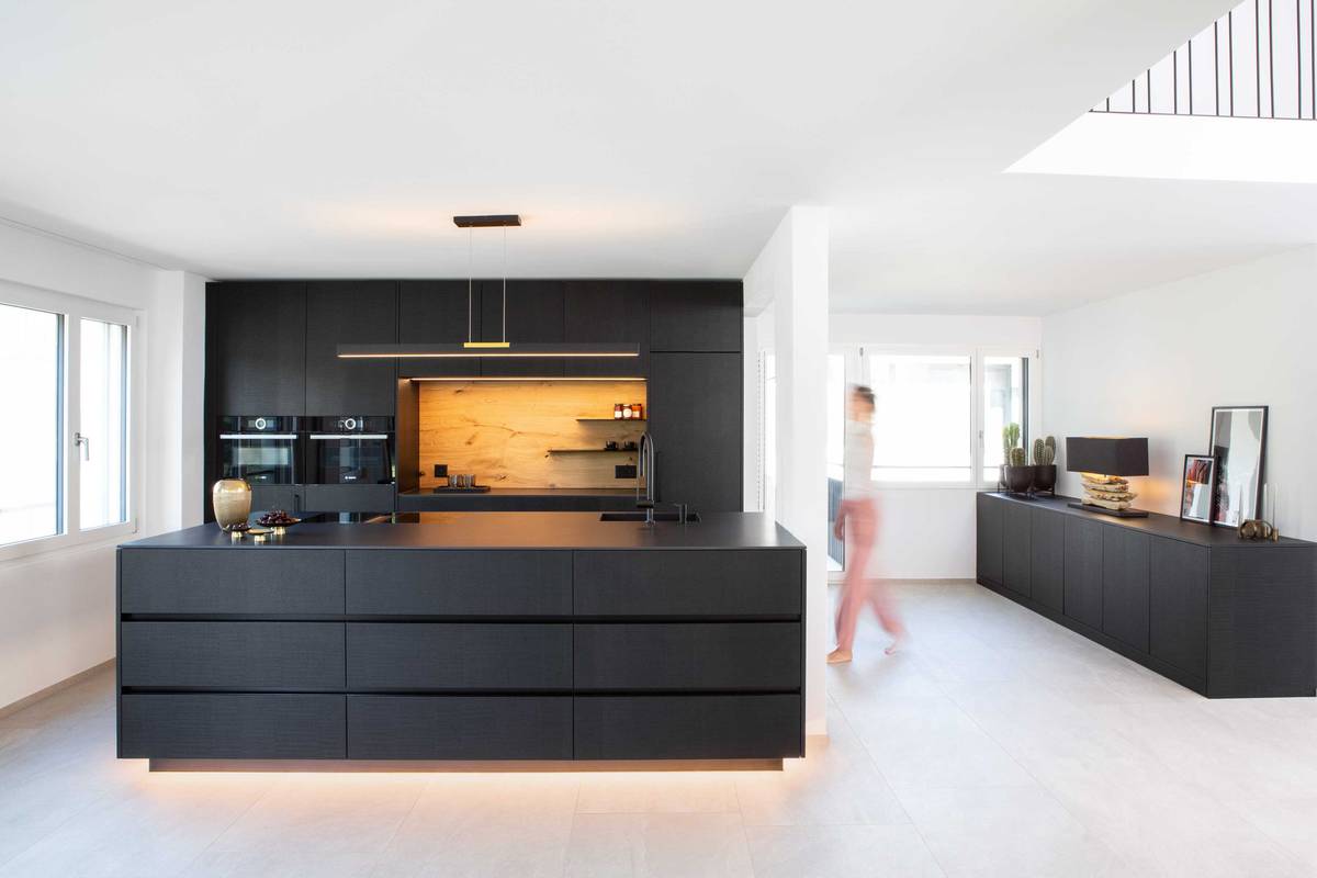 Durch das sägerohe Eichenfurnier, das schwarz gebeizt und lackiert wurde, werden Chalet-Charme und State-of-the-art kombiniert. Durch die Sockelbeleuchtung wirkt die Insel schwebend leicht. Elbau Küchen AG.