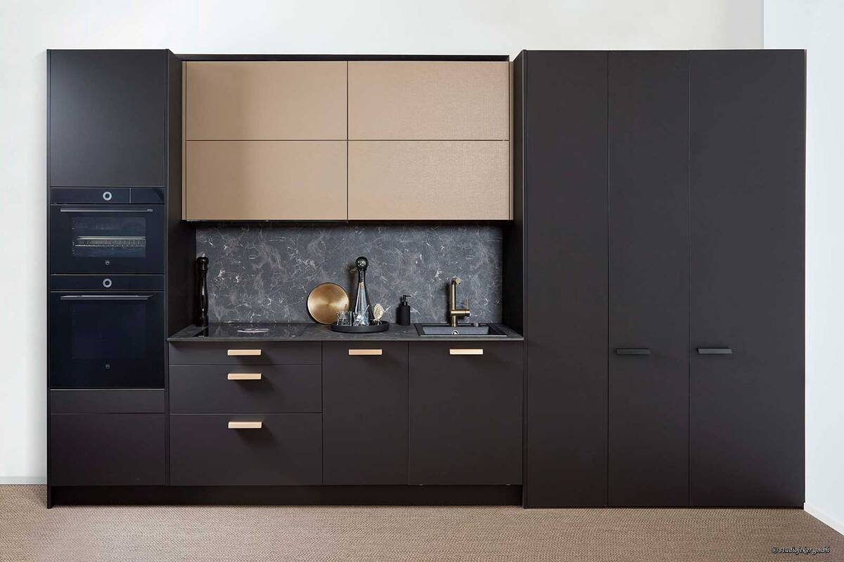 Die mattschwarze Küche mit goldenen Armaturen und Griffen vermittelt Eleganz. Durch die Rückwand und die hellen Oberschränke entsteht am Arbeitsort ein natürlicher Fokuspunkt. Bei der Planung sollte stets berücksichtigt werden, dass wie bei dieser Küche, Backofen und Steamer sich durch das Schwarz optisch in der Küche integrieren und nicht sofort ins Auge springen. Sabag AG.