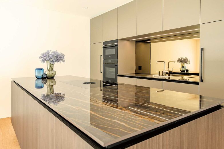 Tiefe und Helligkeit: Diese stilvolle Küche vereint modernes Design mit funktionalen Elementen. Die Fronten in Achatgrau der «X-Line»-Griffleiste wirken elegant und zeitlos. Ein Highlight ist die polierte Keramikabdeckung «Noir Desir», die mit ihrem dezenten Glanz edle Akzente setzt. Eindrucksvoll ist die bronzefarbene Spiegelrückwand, die Licht reflektiert und dem Raum Helligkeit und Tiefe verleiht.
Hans Eisenring AG, www.eisenring-kuechenbau.ch