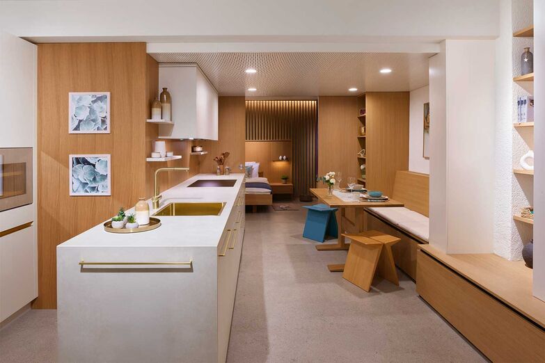 Einrichtung für ein Tiny House: Küche, Essplatz, Stauraum, Schlafzimmer und Bad finden auf kleinstem Raum Platz und bieten ein luftiges und funktionales Ambiente. Die Küche besticht durch einen hellen, warmen Farbton und harmoniert mit der fast gleichfarbigen Keramikabdeckung. Die Griffe, die Mischer und das Becken in zartem Gold sowie die Einbaugeräte und der Weinkühler in der Farbe Pearl verleihen einen Hauch von Luxus. Das Holzfurnier für Wände und Schiebetüre kann in der Produktion und Ausstellung in Wyssachen besichtigt werden. Die zweite Ausstellung befindet sich in Langenthal.
Loosli AG, www.loosli.swiss